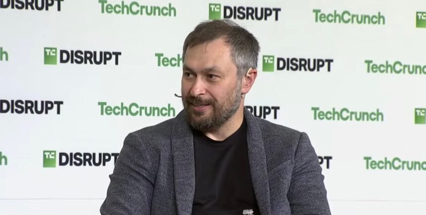 A bearded man grins against a TechCrunch-branded backdrop
